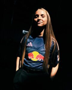 Portrait of a female athlete with long straight hair, wearing a blue Red Bull jersey under a spotlight in a dark setting.