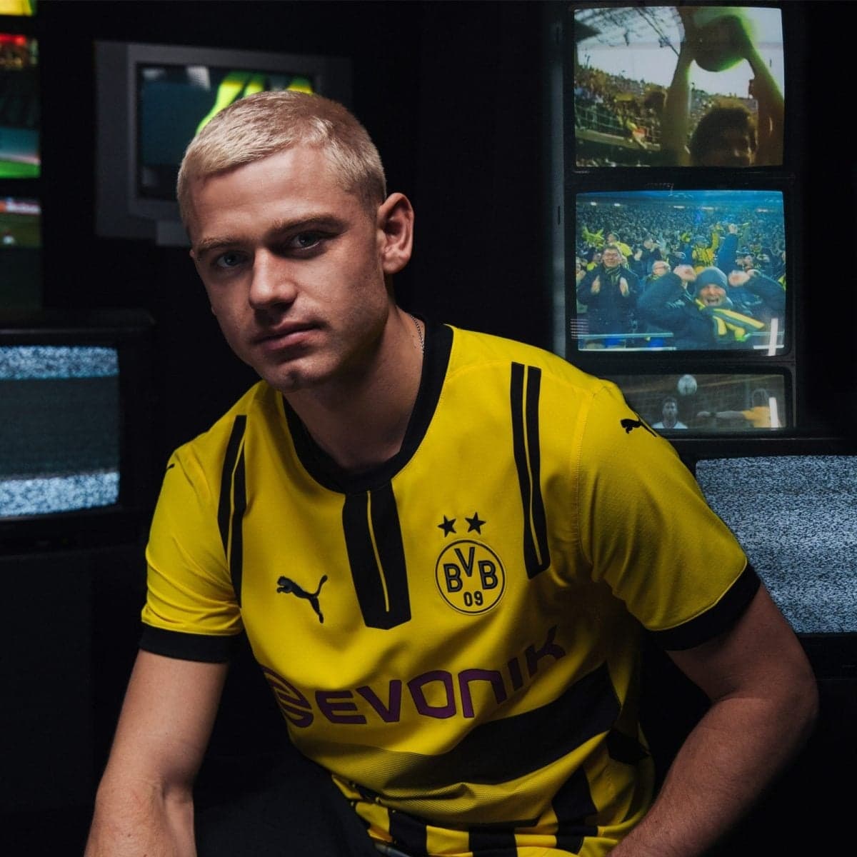Young man with blond hair in a yellow Borussia Dortmund jersey sits in a dark room, with video screens showing fans in the background.