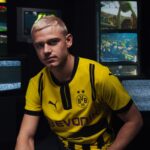 Young man with blond hair in a yellow Borussia Dortmund jersey sits in a dark room, with video screens showing fans in the background.