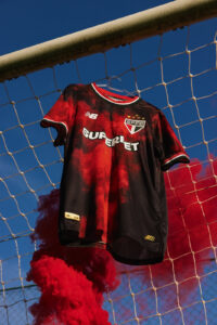 Red and black São Paulo FC soccer jersey with New Balance logo hanging on a goal net against a blue sky, with red smoke at the bottom, and a diagonal white text logo across the chest.