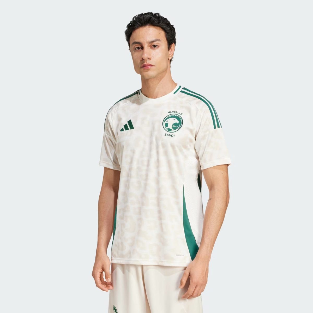 Young man wearing a white Saudi football jersey with green stripes and crest, standing against a light background.