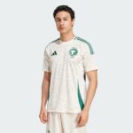 Young man wearing a white Saudi football jersey with green stripes and crest, standing against a light background.