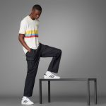 Man in a white striped t-shirt and black cargo pants steps onto a metal table in a studio with a gray backdrop, one foot on the edge.