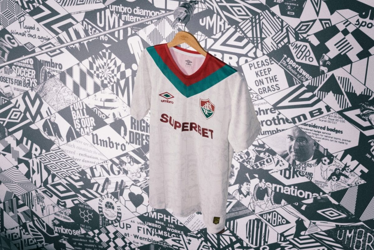 White football jersey on a hanger in front of a collage wall; features a green and red V-neck, Umbro logo, club crest, and the sponsor text 'SUPERBET'
