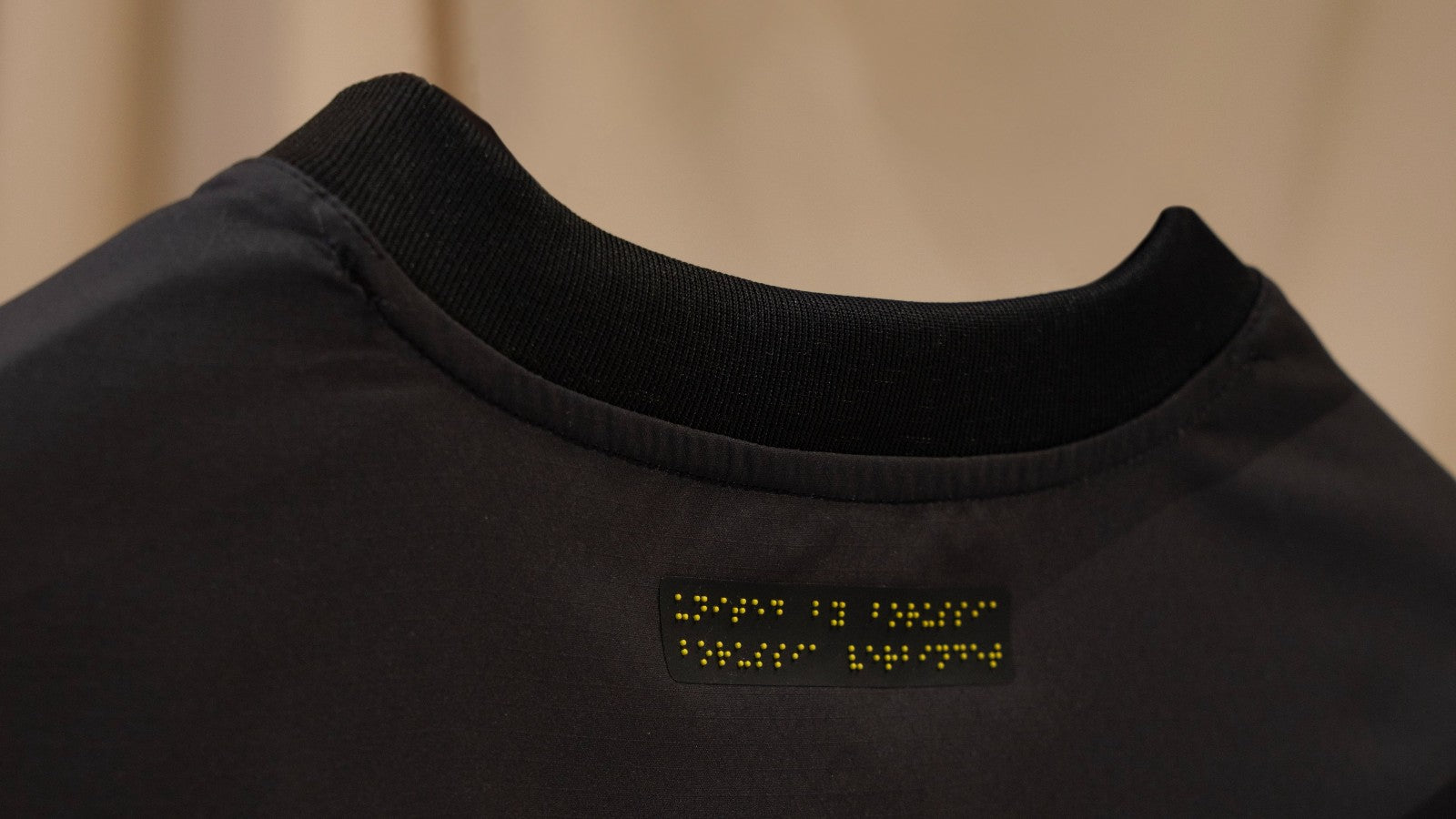 r5MoHEsCZjm5qOs | UAE SPORT STORE Close-up of black shirt neckline with a small black patch featuring yellow Braille text on the back.