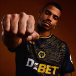 Man with a serious expression extends a clenched fist toward the camera, wearing a dark jersey with yellow detailing.
