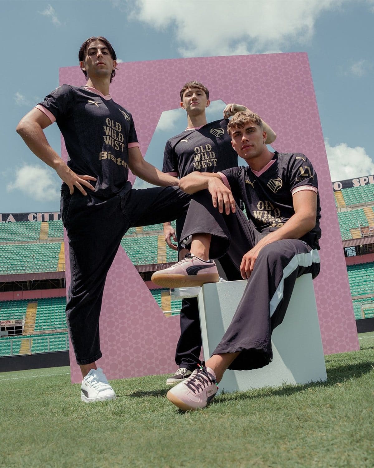 Three men in matching black-and-pink shirts pose on a football field, with a large pink patterned frame behind them.