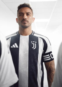 Male soccer player in a black-and-white Juventus jersey stands in a bright corridor, looking ahead with a serious expression.