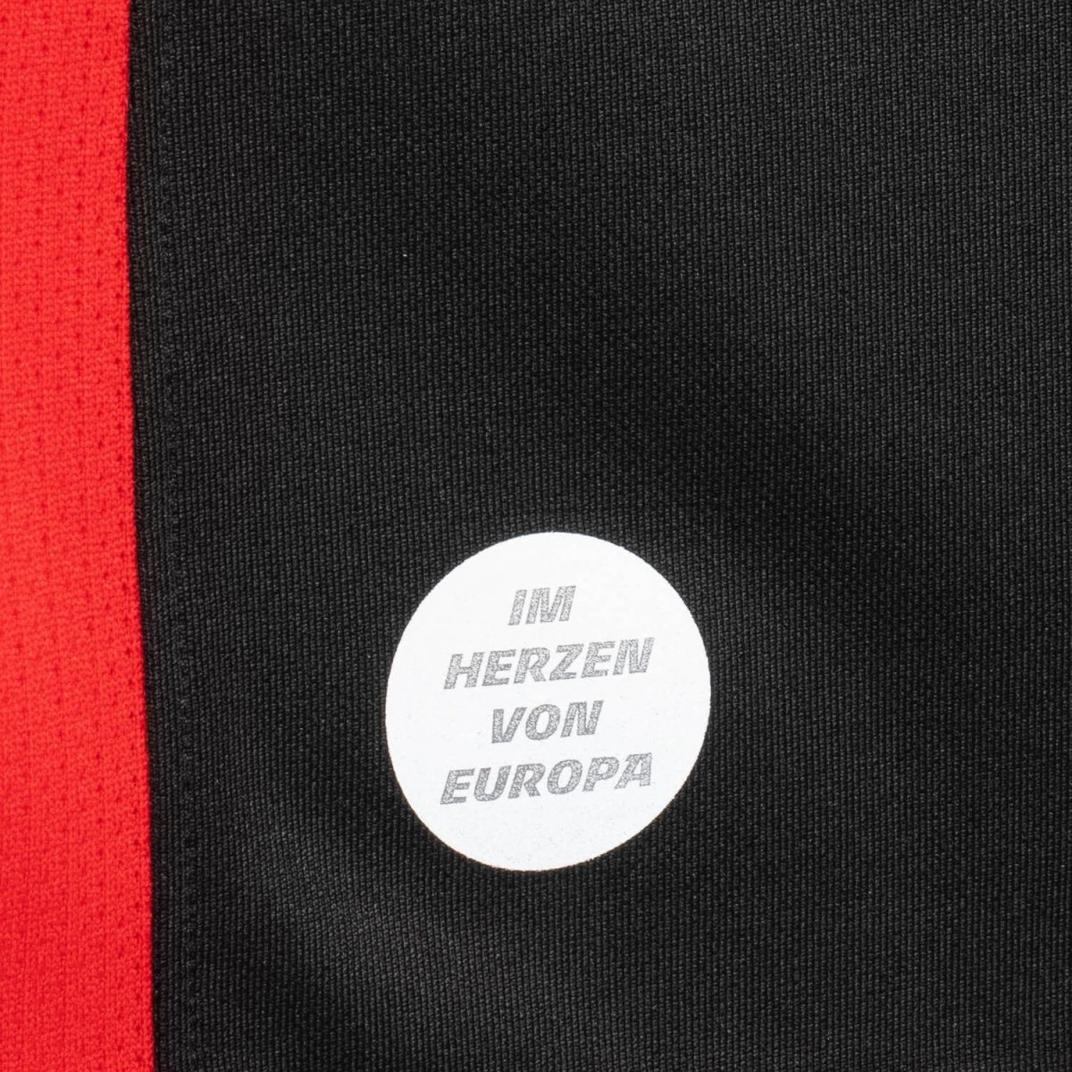 Close-up of black knit fabric with a vertical red stripe on the left and a white circular sticker that reads "IM HERZEN VON EUROPA".