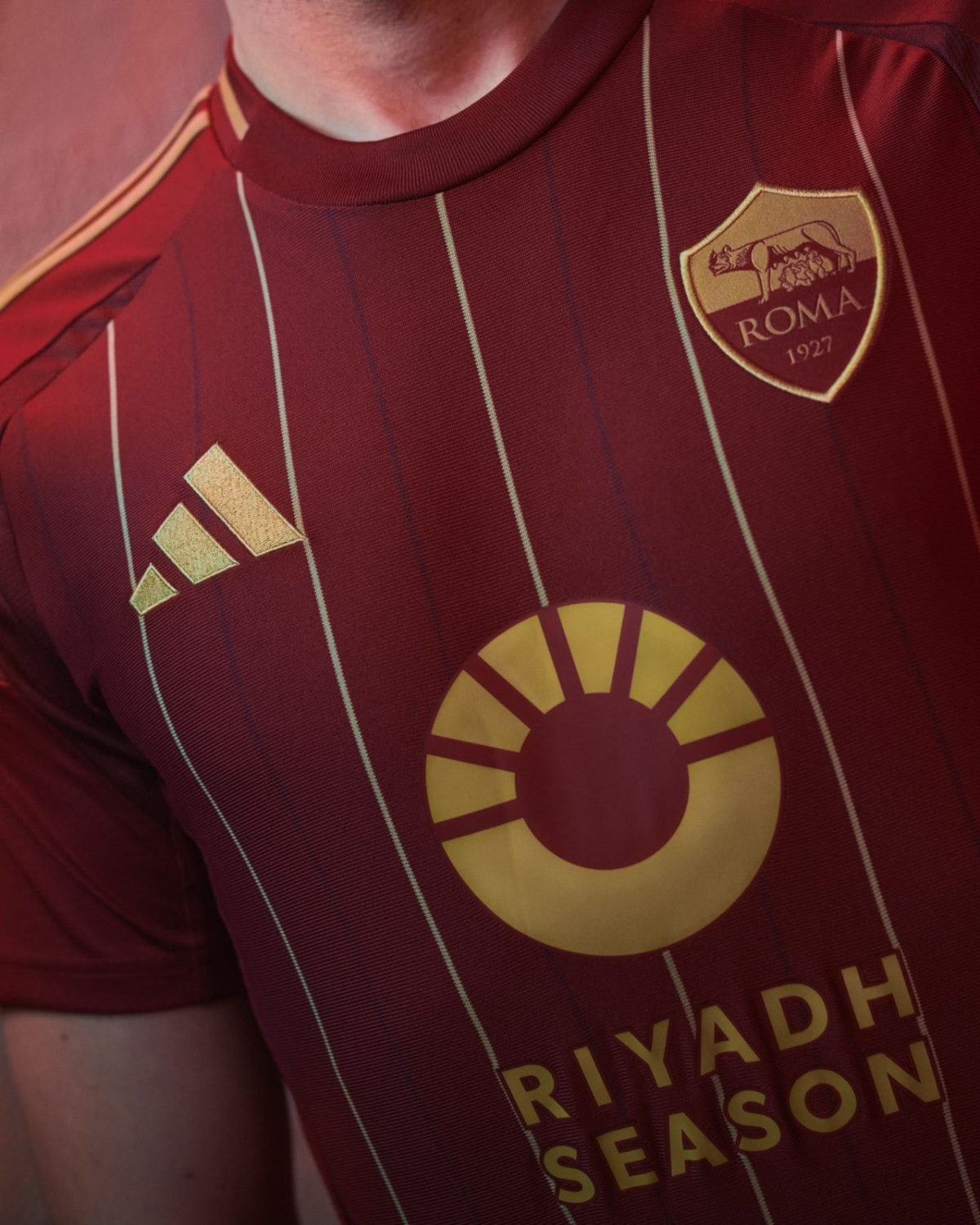 Close-up of a maroon Roma football jersey with vertical white stripes, Adidas logo, and the Roma crest (1927).
