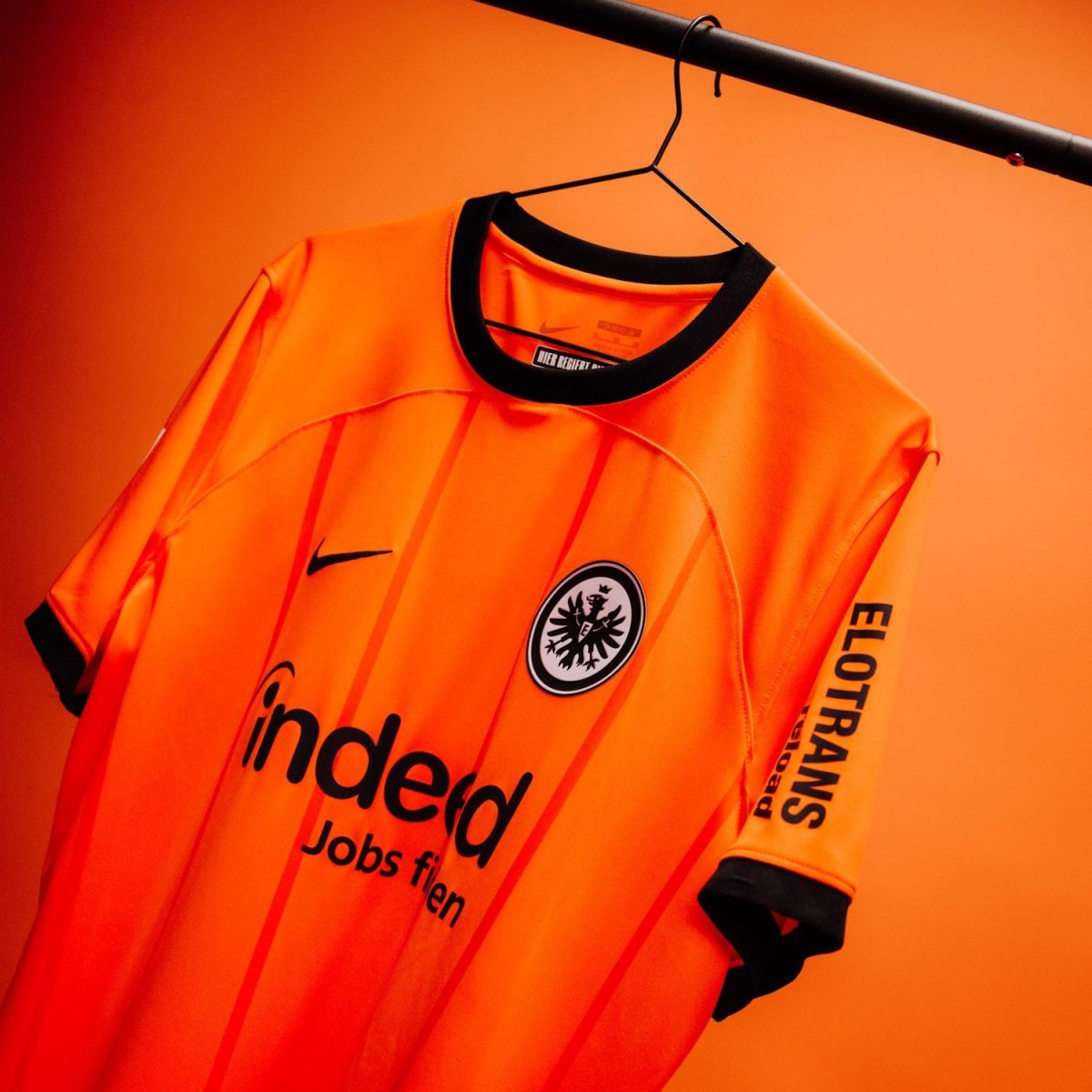 Orange Eintracht Frankfurt football jersey hanging on a black hanger against an orange backdrop, showing the club crest and sponsor Indeed.