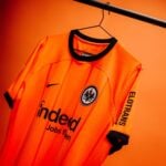 rWsUgM5W4QrIPUV | UAE SPORT STORE Orange Eintracht Frankfurt football jersey hanging on a black hanger against an orange backdrop, showing the club crest and sponsor Indeed.