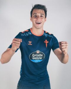 Young man in a blue sports jersey raises his fists in celebration, smiling at the camera.