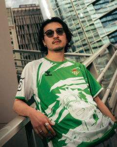 Man with shoulder-length dark hair and round sunglasses leans on a railing, wearing a green-and-white Naruto-themed jersey in a city setting on a sunny day.