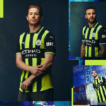 Portrait of a man in a neon-yellow and navy Manchester City jersey, arms crossed, against a dark backdrop.