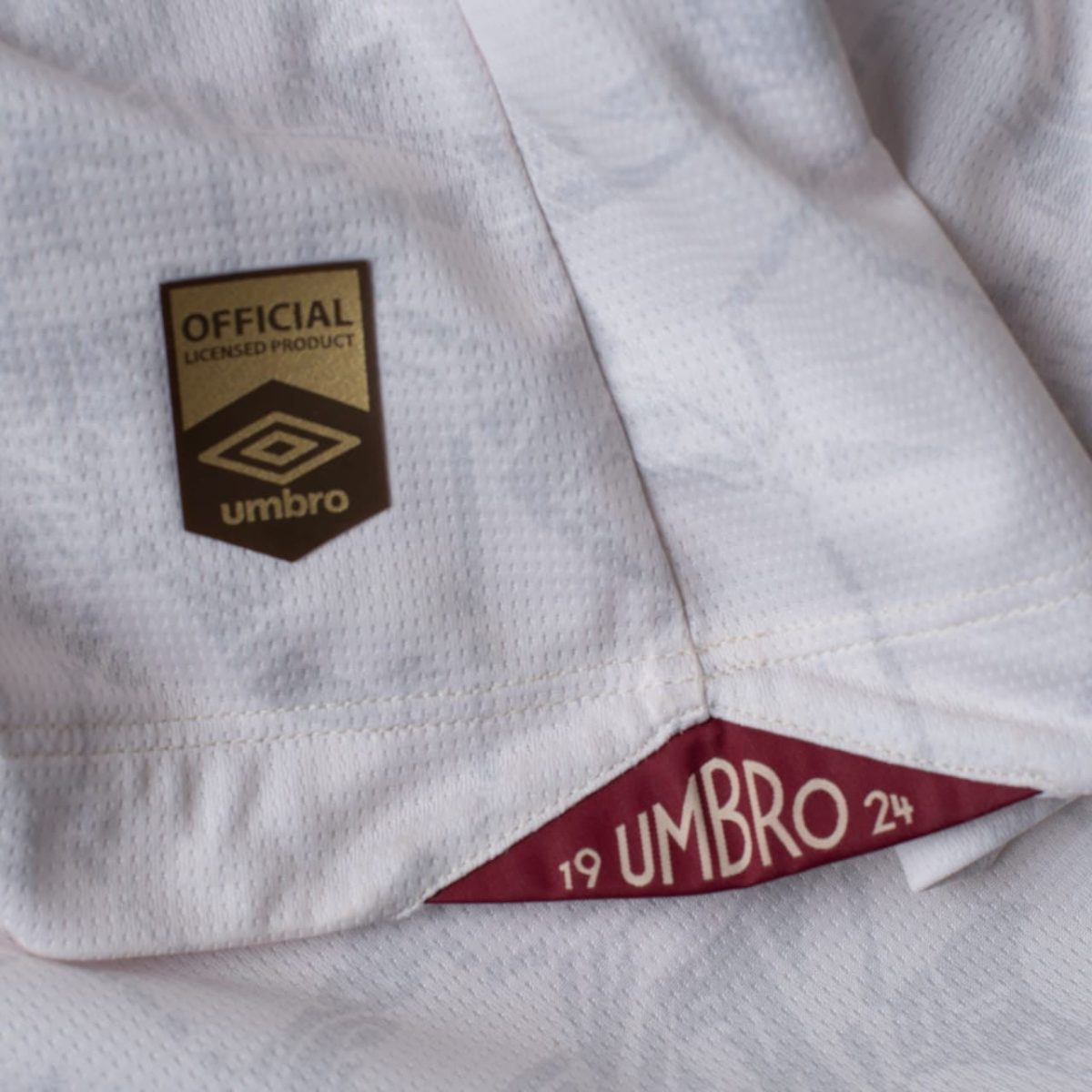 Close-up of a white athletic jersey showing an official licensed product badge with the Umbro logo on a gold-and-black patch alongside a red label edge with 'UMBRO' text.