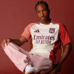 Portrait of a man seated on a chair, wearing an OL Lyonnais football jersey with the 'Emirates Fly Better' slogan and light pink sweatpants against a red backdrop.