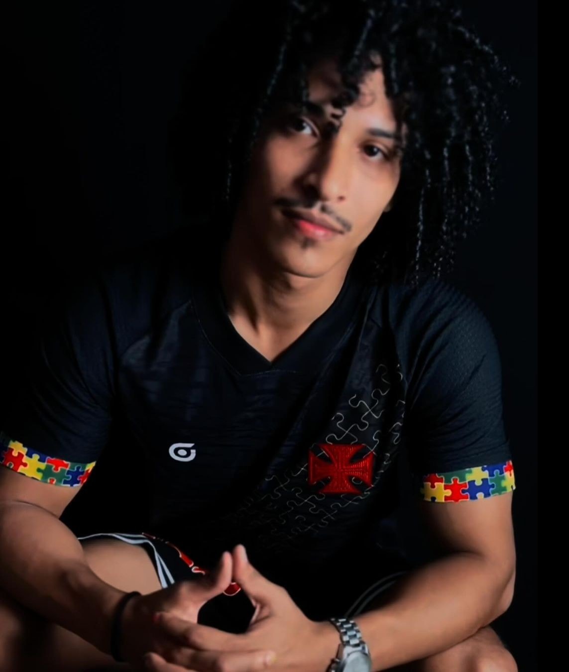 rmdqB1zPzdds2GU_1 | UAE SPORT STORE Portrait of a young man with curly hair and a slight smile, wearing a black shirt with colorful puzzle-piece sleeves, hands clasped in front against a dark background.