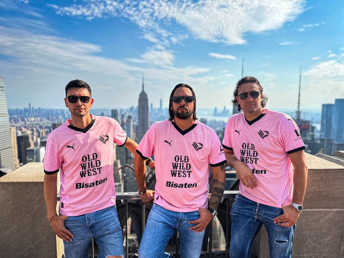 Three men in pink jerseys pose on a rooftop with a New York City skyline in the background.