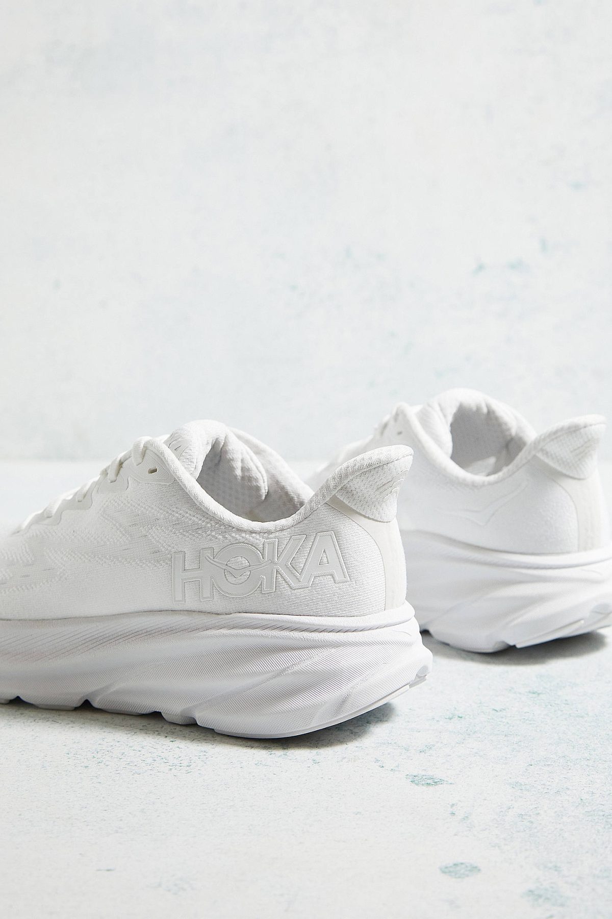 White HOKA running shoes photographed close-up on a pale, textured surface. More focus on the front shoe with logo visible on the side.