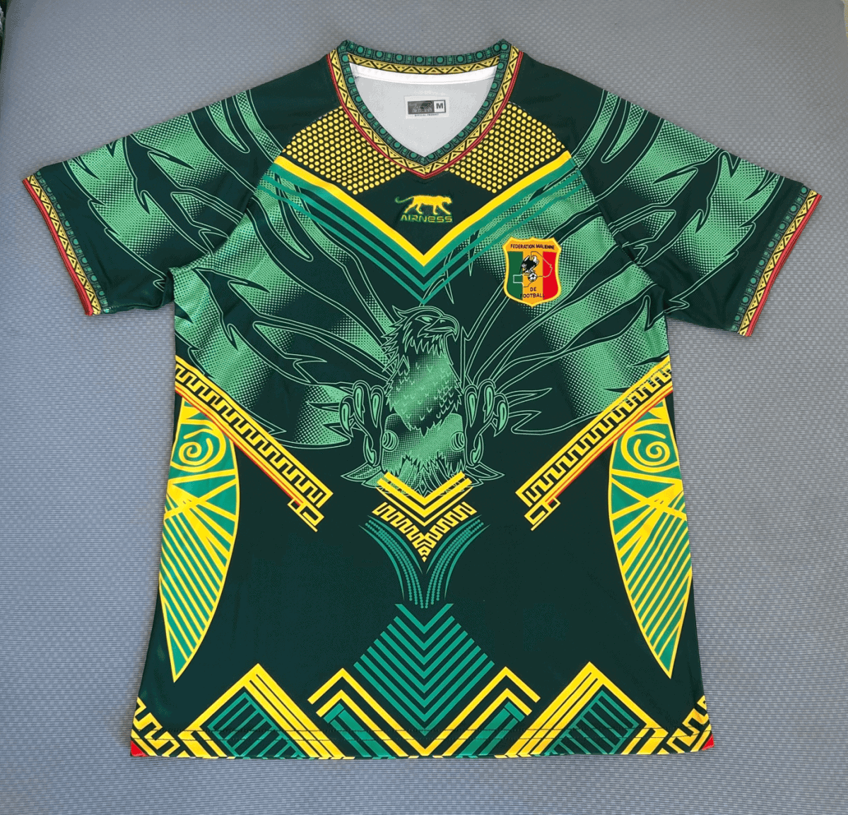 Green football jersey with yellow and teal geometric patterns and a shield crest reading Federation Malienne de Football on the chest, featuring an eagle motif.