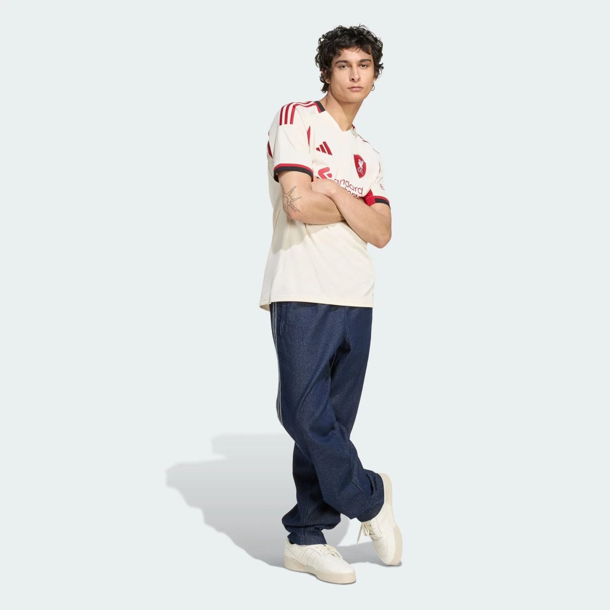 Young man posing in a white jersey with red accents and blue jeans against a pale background, white sneakers on.