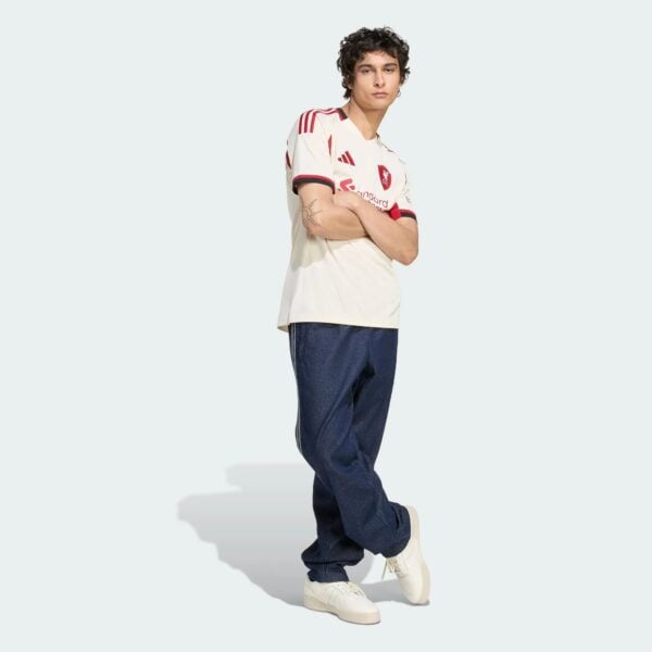Young man posing in a white jersey with red accents and blue jeans against a pale background, white sneakers on.