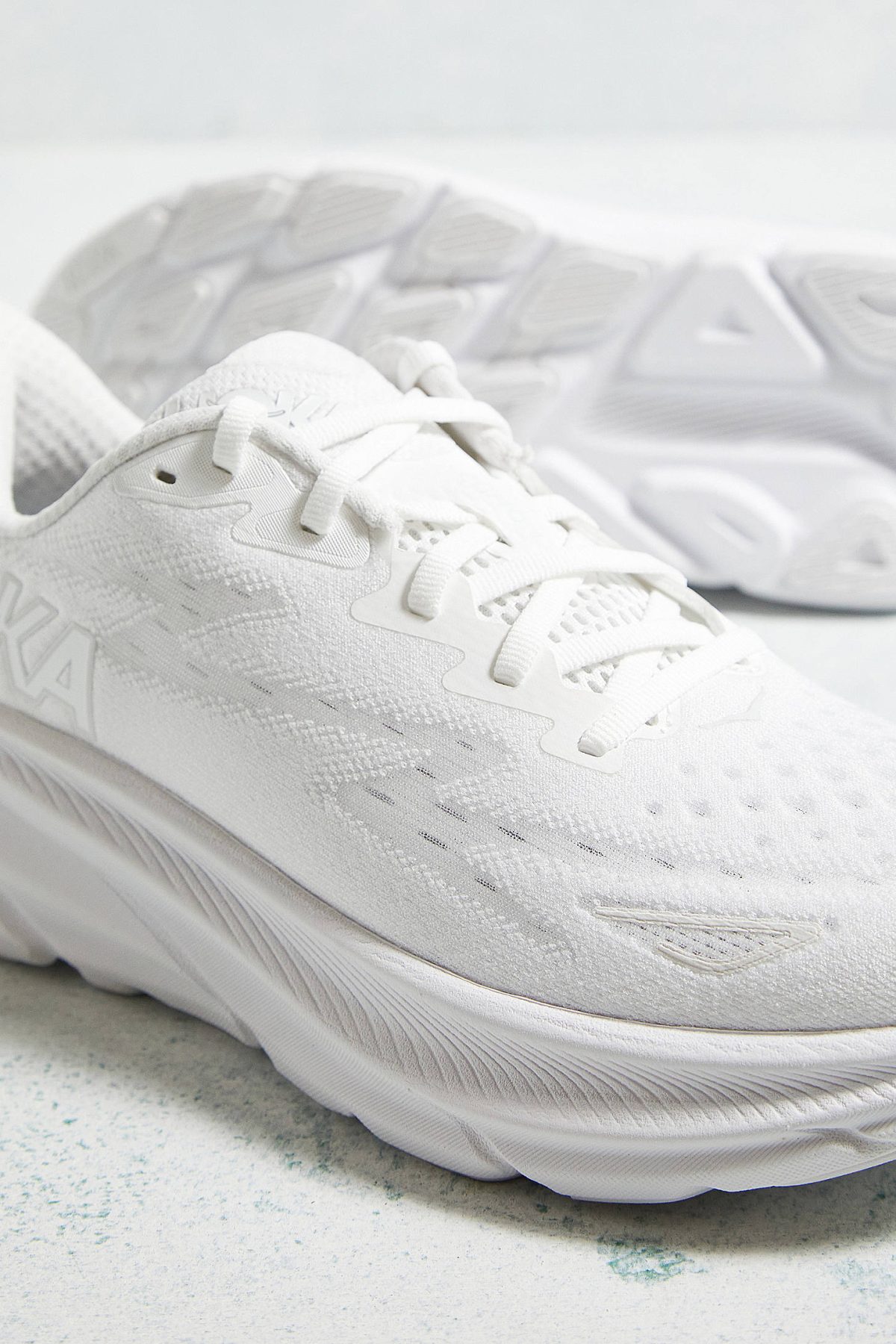 Close-up of white running shoes with laces, knit texture, and a thick sole on a light speckled surface.