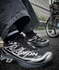 rn-image_picker_lib_temp_0672ff1c-7c05-41d4-a6cc-2b614edf3683 | UAE SPORT STORE Close-up of a person's black and silver athletic shoes with geometric panels, wearing cuffed jeans and striped socks, on asphalt beside a bicycle.