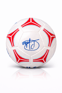 White soccer ball with red geometric panels and a blue signature-style logo, on a white reflective surface, front view to highlight design.
