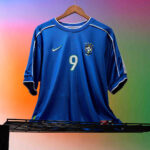 rn-image_picker_lib_temp_0966e865-a892-40d7-bd5f-e75fc61f82c7 | UAE SPORT STORE Blue Brazil national team soccer jersey with number 9 hanging on a wooden hanger against a colorful gradient background.