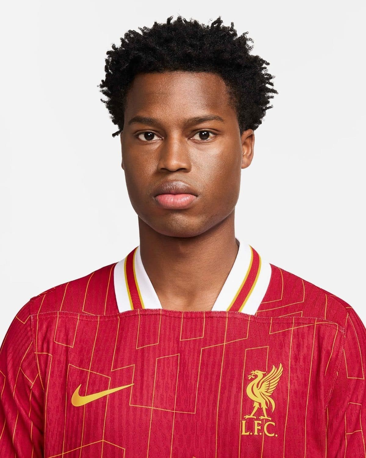 Close-up portrait of a young man with short curly hair wearing a red Liverpool FC football jersey with a gold crest and yellow Nike swoosh, looking at the camera.