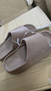 Brown slide sandals with textured footbeds resting on a cardboard box. A plastic security tag loop sits on one sandal.