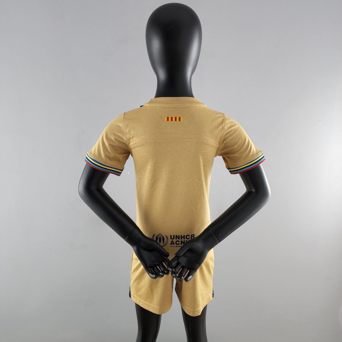 Back view of a black mannequin wearing a tan short-sleeve shirt with striped sleeve cuffs and UNHCR logo on the lower back, arms crossed behind the torso.