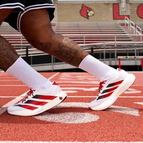 Athlete wearing white and red track shoes sprinting on a red running track at a stadium.