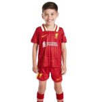 Front view of a smiling child wearing a red Liverpool FC kids football kit with sponsor logos, standing against a white background.