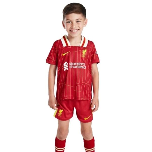 Front view of a smiling child wearing a red Liverpool FC kids football kit with sponsor logos, standing against a white background.