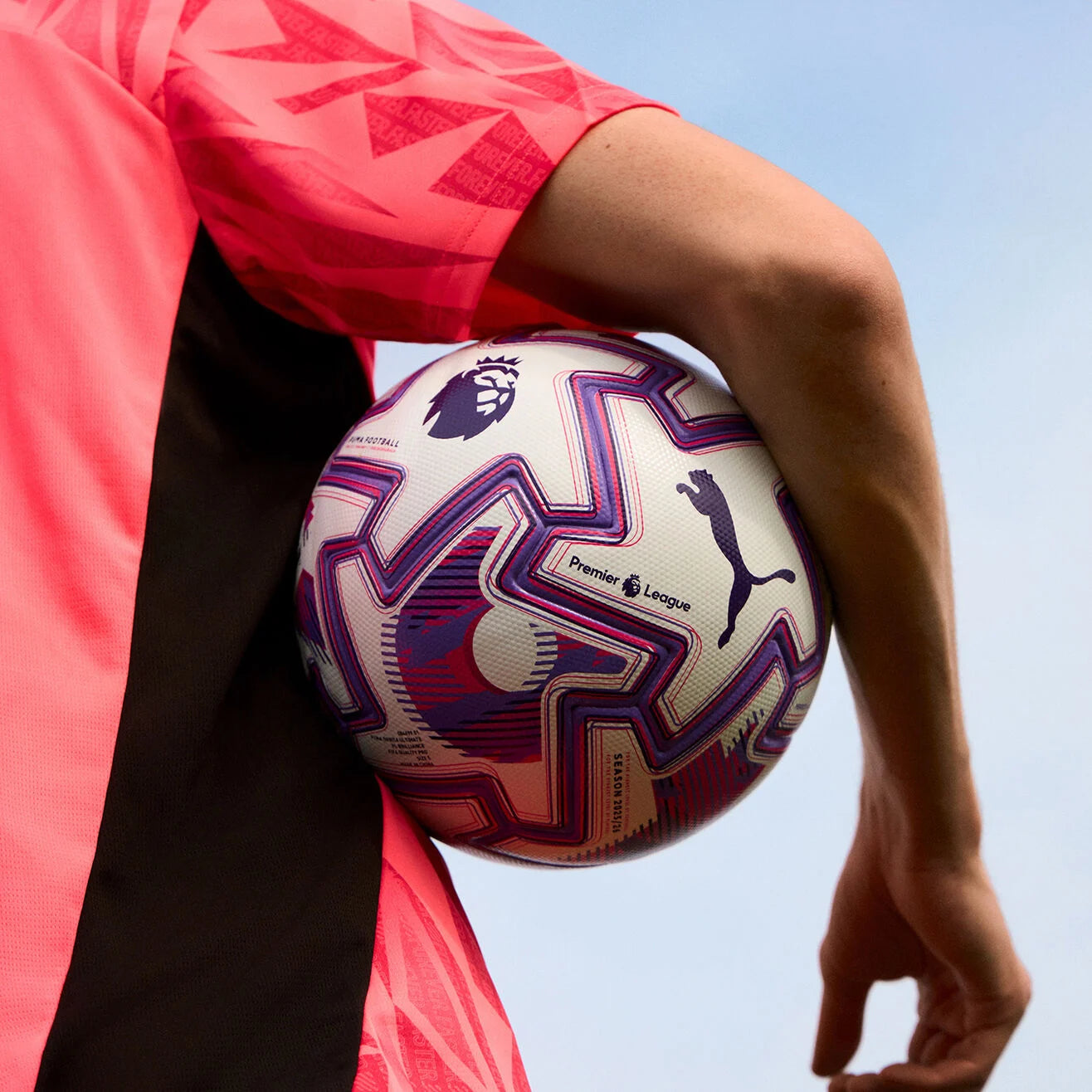 rn-image_picker_lib_temp_1d940155-dbd7-48cb-b35c-76e3a4060b65 | UAE SPORT STORE Soccer player in a pink jersey holding a white Premier League ball under their arm against a light blue sky.