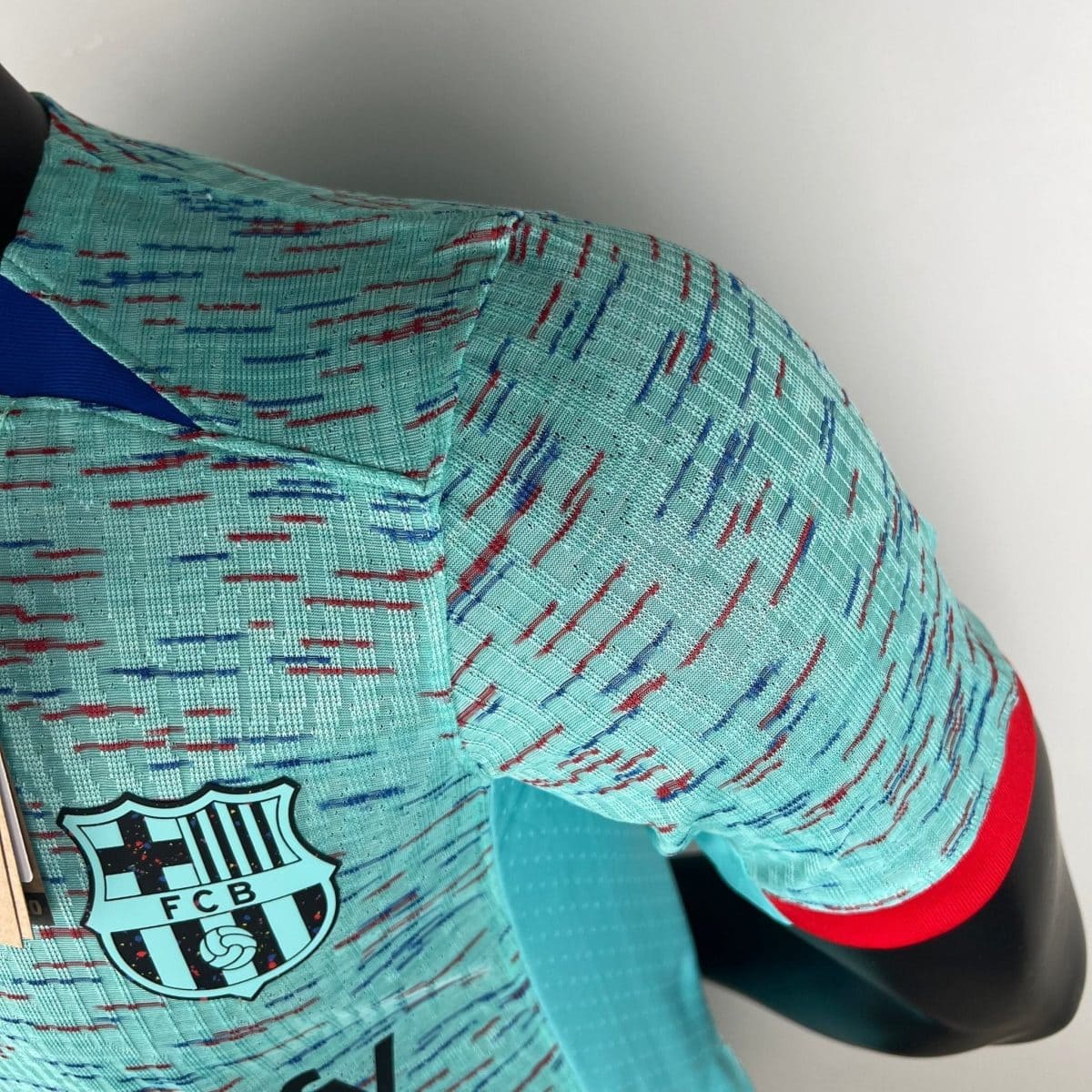 Close-up of turquoise sports jersey fabric with red and blue stitching, showing a partial FC Barcelona crest.