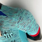 Close-up of turquoise sports jersey fabric with red and blue stitching, showing a partial FC Barcelona crest.