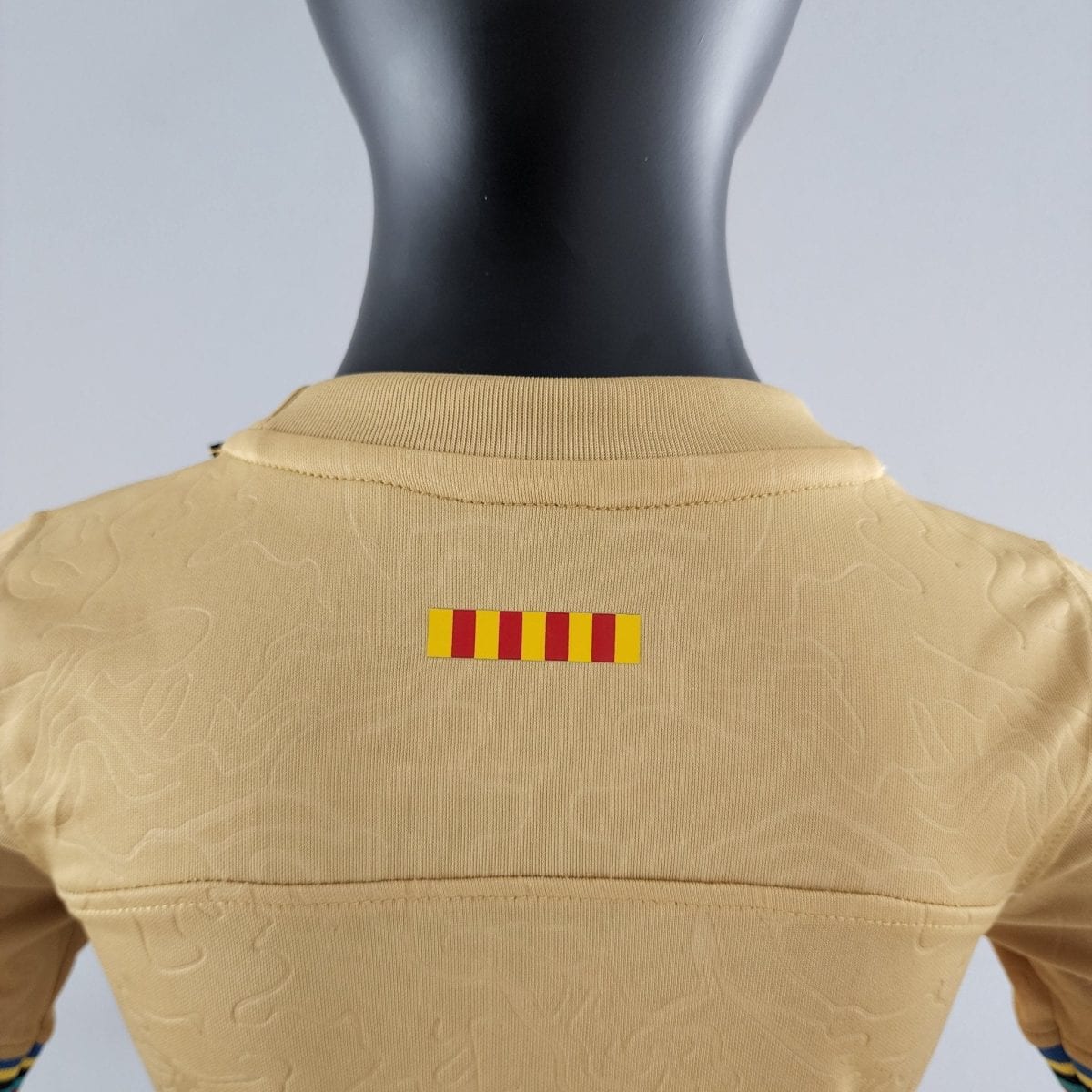 Back view of a black mannequin wearing a tan knit top with a small yellow-and-red striped label on the chest area.