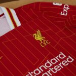 Red Liverpool FC jersey with yellow crest and geometric orange lines, white sponsor text visible at the bottom edge, collar detail nearby.