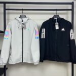 rn-image_picker_lib_temp_2a138eeb-f744-42da-aed0-be29d46ab03f | UAE SPORT STORE Two Adidas windbreaker jackets on a clothing rack: left light gray with iridescent sleeve stripes and small colorful logo; right black with white Adidas stripes on the sleeves and a white logo.
