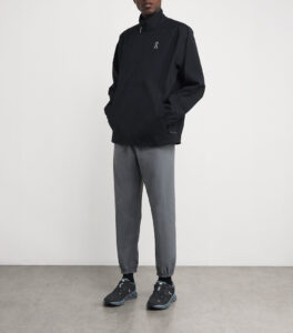 rn-image_picker_lib_temp_36730176-0114-4901-9cd9-9d644826c25a | UAE SPORT STORE Person wearing a black windbreaker with a small white logo, gray tapered jogger pants, and black hiking-style sneakers, standing indoors.