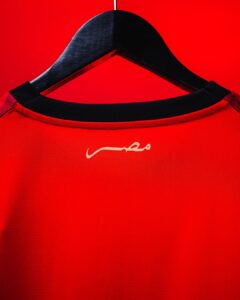 Red athletic jersey on a black hanger against a red background, with a small white cursive logo near the collar on the back of the shirt.