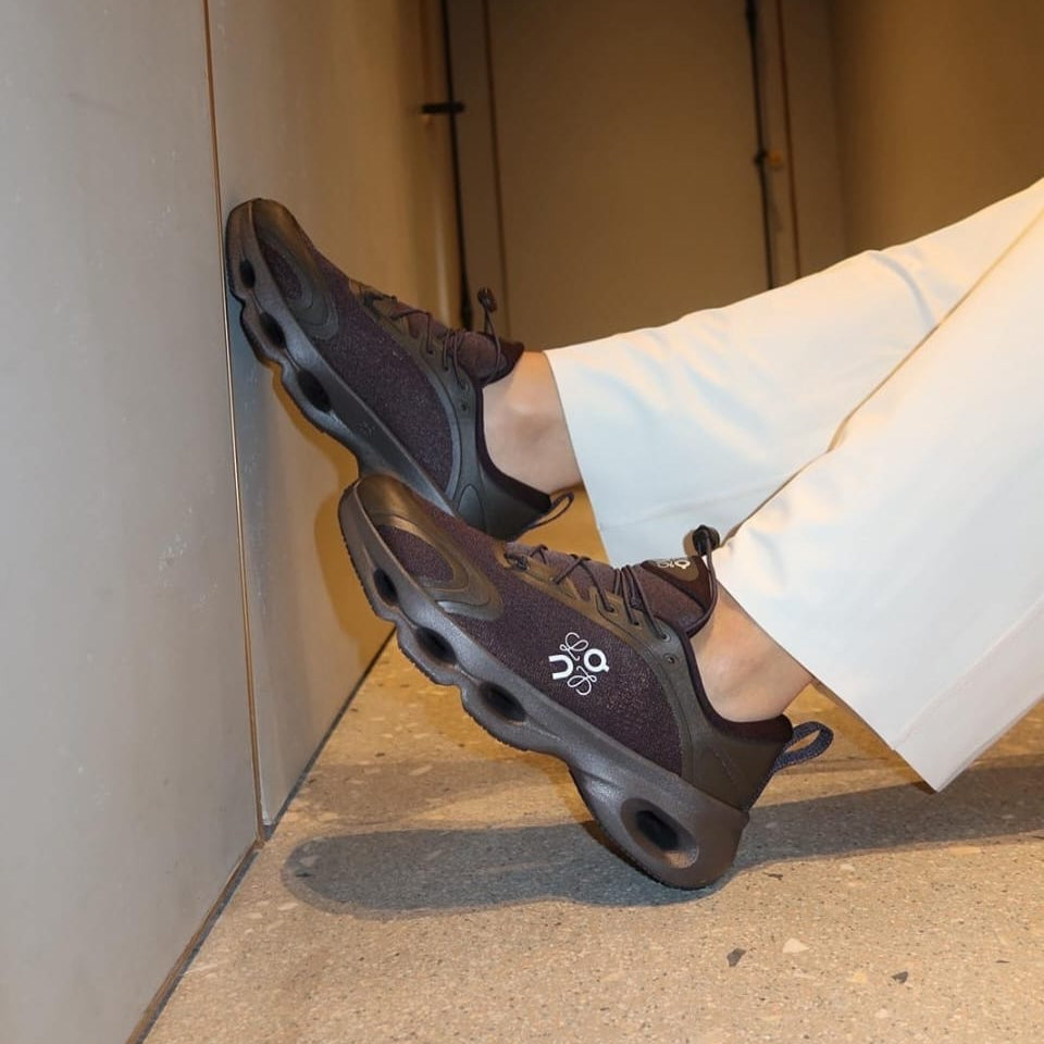rn-image_picker_lib_temp_4947316e-d84a-40a7-9b92-852d3fd1b408 Person wearing dark purple athletic sneakers with a chunky black sole, feet propped against a wall in white pants.