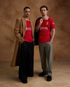 Two men stand side by side in red Liverpool FC jerseys against a brown studio backdrop; one wears a camel overcoat.