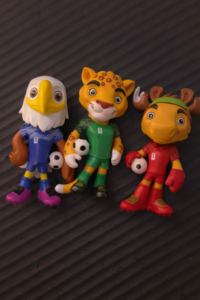 Three toy soccer players: a white bird in blue, a leopard in green, and a moose in red, each with a small soccer ball.