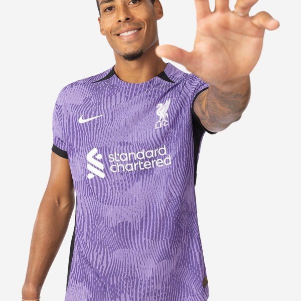 Man in a purple Liverpool FC jersey smiles and reaches his hand toward the camera.