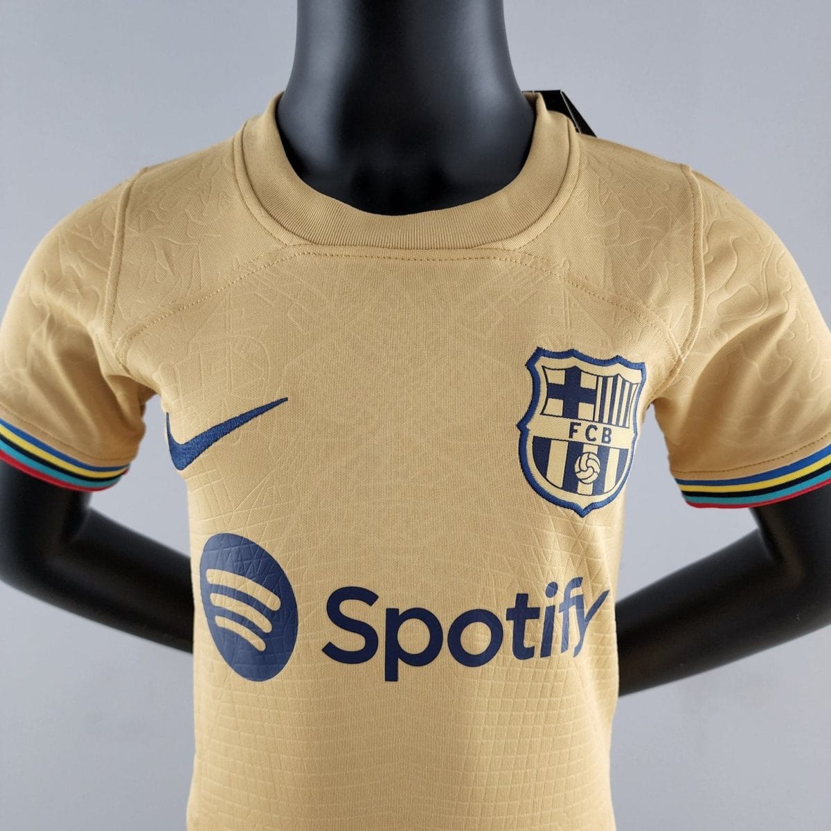 Close-up of a beige sports jersey on a mannequin, showing a blue Nike swoosh and a crest with FCB on the chest, plus striped sleeve edges.
